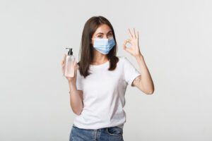 covid 19 health and social distancing concept portrait of smiling brunette girl in medical mask showing hand sanitizer and okay gesture